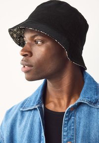 Cappello a secchiello nero con interno fantasia. Presenta una tesa larga e un tessuto strutturato. Indossato con una giacca di jeans e una maglietta nera.