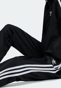 Černý sportovní teplákový komplet vyrobený z hladkého materiálu, s bílými třemi pruhy po stranách a malým logem na kalhotách.