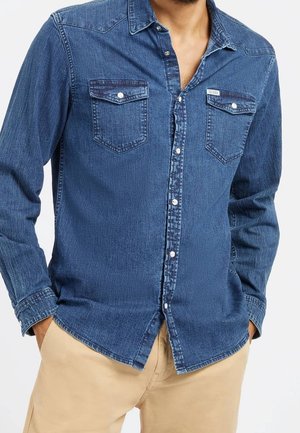 Homme portant une chemise en denim bleu boutonnée avec deux poches poitrine et un pantalon beige, les mains dans les poches, sur un fond uni.