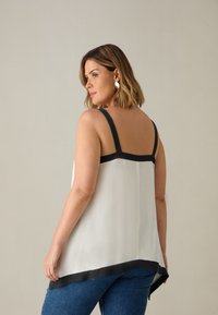Top bianco senza maniche con bordatura nera, caratterizzato da spalline larghe e una vestibilità ampia. Il retro è leggermente più lungo, creando un effetto sovrapposto.