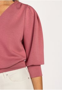 Roze top met korte pofmouwen, een gladde textuur en een v-hals ontwerp. Gecombineerd met lichtgekleurde broeken.