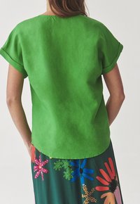 Top verde a maniche corte in un tessuto liscio, caratterizzato da un collo rotondo e un orlo leggermente curvo, abbinato a una gonna con motivo floreale multicolore.