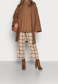 Femme portant un manteau en laine marron, un pull en maille torsadée, un pantalon écossais court, des bottines marron et un sac bandoulière marron.