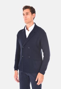 Cardigan lavorato a maglia blu navy con design doppiopetto, grandi bottoni e tasche laterali. La texture è a coste, con un colletto a scialle.