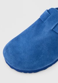 Γαλάζιο σουέντ παντόφλα με στρογγυλερή μύτη και παχιά σόλα σε ανοιχτό γκρι φόντο.