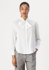 Camicia bianca a maniche lunghe con colletto a fiocco, realizzata in tessuto morbido. Abbinata a pantaloni grigi a quadri, con una vestibilità morbida.