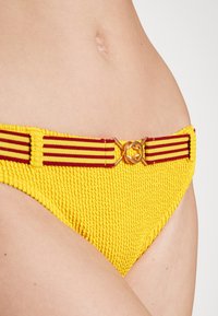 Parte de baixo de biquíni amarela com tecido texturizado, apresentando uma cintura larga em listras vermelhas e detalhe em metal dourado no centro.