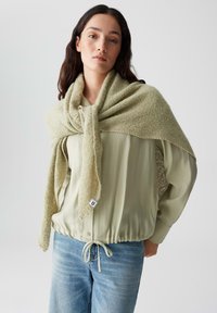 Lichtgroene sjaal van textuurstof, losjes over een crèmekleurige top met een gathered tailleband gedrapeerd, gecombineerd met lichtblauwe jeans.