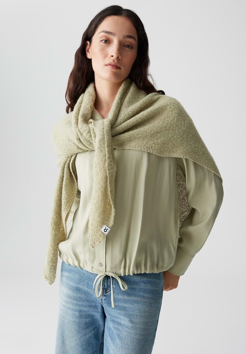 Lichtgroene sjaal van textuurstof, losjes over een crèmekleurige top met een gathered tailleband gedrapeerd, gecombineerd met lichtblauwe jeans.