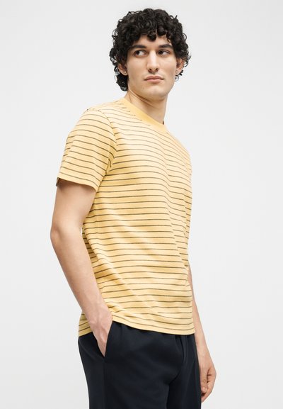 Νεαρός με σγουρά μαλλιά, ντυμένος με κίτρινο κοντομάνικο πουκάμισο με λεπτές οριζόντιες ρίγες και σκούρο παντελόνι, με τα χέρια στις τσέπες, κοιτάζει πλάγια.