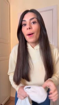 Femme aux longs cheveux bruns portant un sweat à capuche clair, tenant un vêtement blanc plié à l'intérieur, près d'une porte blanche et d'une armoire.