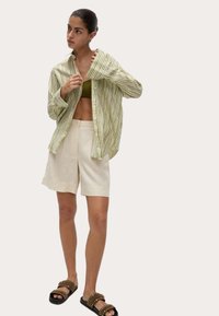 Camicia a righe verde e bianca con bottoni, pantaloni corti beige e sandali scuri con borchie. La camicia è aperta, rivelando una canotta oliva sotto.