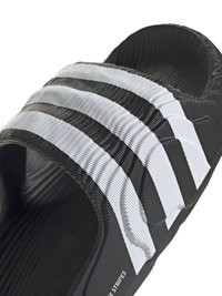 Crne klizne sandale s tri istaknute bijele pruge preko gornjeg dijela. Teksturirani gumeni materijal, otvorena peta i strukturni dizajn uloška.