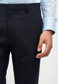 Mornarske krojene hlače od glatkog materijala s ravnim prednjim dijelom, petljama za remen i uskim krojem. Ruka se naslanja na bočni šav.