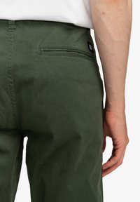 Pantaloni in cotone verdi con tasca posteriore, dotati di un bottone con logo nero circolare. Tessuto liscio e vestibilità su misura.