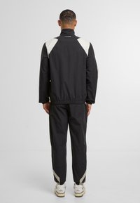 Giacca sportiva nera con accenti bianchi sulle spalle, polsini elastici e pantaloni coordinati. Indossato con scarpe di colore neutro con marchio.
