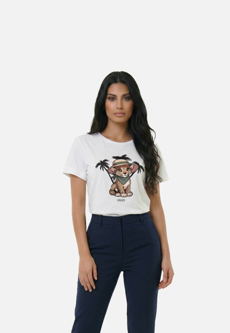 Femme aux longs cheveux noirs portant un t-shirt blanc avec un léopardon coiffé d'un chapeau et entouré de palmiers, associé à un pantalon bleu foncé.