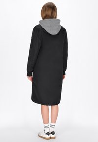 Abrigo acolchado negro con capucha gris, mangas largas, bolsillos laterales y puños acanalados, que llega hasta la mitad de la pantorrilla, combinado con zapatillas blancas y calcetines a rayas.