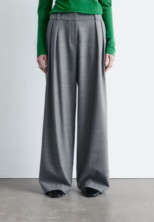 Graue, hochtaillierte, plissierte Hose mit weitem Bein, aus strukturiertem Stoff, kombiniert mit einem eng anliegenden grünen Oberteil und schwarzen Schuhen.