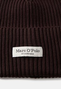 Gorro de punto acanalado marrón que presenta una etiqueta blanca con texto negro que dice "Marc O'Polo EST. EN ESTOCOLMO." Texturizado con detalles visibles de hilo.