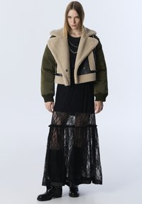 Frau in olivgrünem Schaffelljacke über langem schwarzem Spitzenkleid mit schwarzen Stiefeln, stehend vor einem schlichten hellen Hintergrund.