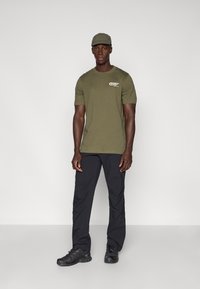 Camiseta de manga corta verde oliva con logo, pantalones negros y zapatillas deportivas negras. Diseño simple, corte regular, tejido ligero, combinado con una gorra a juego.