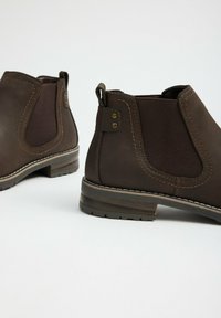 Par de botas Chelsea de cuero marrón con paneles laterales elásticos, tiradores y tacones bajos de madera sobre fondo blanco.