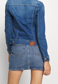 Jeansowa kurtka o teksturowanym niebieskim wykończeniu, z dopasowanym krojem i zapięciem na napy. Połączona z pasującą spódnicą dżinsową, obie mają widoczne szwy.