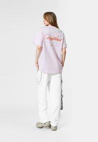 Camiseta de gran tamaño en color rosa claro con texto naranja "Unfettered" en la parte posterior, combinada con pantalones blancos y calzado gris, mostrando un look casual.