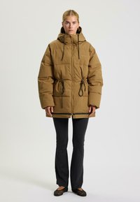 Bruine gewatteerde jas met een capuchon, voorzien van elastische trekkoorden in de taille en aan de onderkant, een ritssluiting aan de voorkant en een ontspannen fit voor comfort.