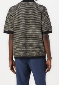Eine Person trägt ein kurzärmliges Schwarz- und Beigetönige gestreiftes Shirt, das mit blauen Hosen kombiniert ist, und ist von hinten vor einem weißen Hintergrund zu sehen.