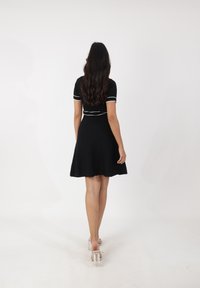 Schwarzes kurzärmliges Kleid mit weißer Borte, weit ausgestelltem Rock und figurbetonter Oberseite. Tragen mit transparenten Absatzschuhen, von hinten betrachtet.