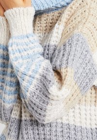 Tjock stickad tröja med breda ränder i krämfärg, grått, beige och ljusblått, som visar en texturerad ribbning och en vikt arm över bröstet.