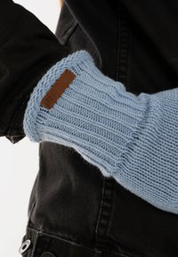 Des gants tricotés bleu clair présentent un design à côtes et une étiquette en cuir marron. Portés par-dessus une veste noire avec des boutons visibles.
