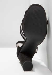 Escarpin en daim noir à talons hauts avec une semelle lisse et brillante, un design à bout ouvert et des détails de brides croisées sur le dessus.
