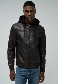 Zwarte leren jack met een ritssluiting aan de voorkant, een capuchon kraag en geribbelde manchetten. Draagt over blauwe denim jeans, met gestikte details.