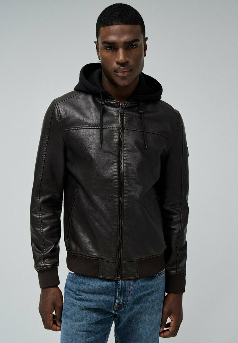 Zwarte leren jack met een ritssluiting aan de voorkant, een capuchon kraag en geribbelde manchetten. Draagt over blauwe denim jeans, met gestikte details.