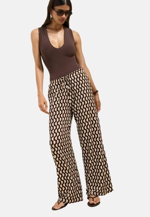 Žena oblečená v hnedom tielku bez rukávov, široké nohavice s béžovo-hnedým vlnkovaným vzorom, slnečné okuliare, náušnice a sandále, stojaca na bielom pozadí.