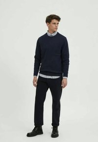 Ναυτικό πλεκτό πουλόβερ πάνω από ένα ανοιχτό μπλε πουκάμισο με γιακά, συνδυασμένο με σκούρα παντελόνια και μαύρα παπούτσια. Απλός σχεδιασμός, λεία υφή και κομψή εφαρμογή.