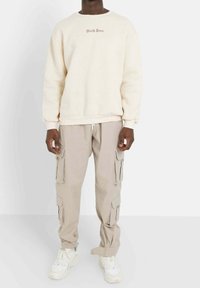Crèmekleurige sweatshirt met "Sixth June" borduursel, gecombineerd met beige cargo pants met meerdere zakken en elastische boorden. Witte sneakers.