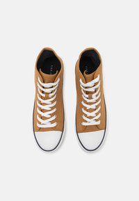 Høye sneakers i tan lerret med hvite snørebånd, hvit gummitåhette og kontrasterende marineblå kant langs sålen.