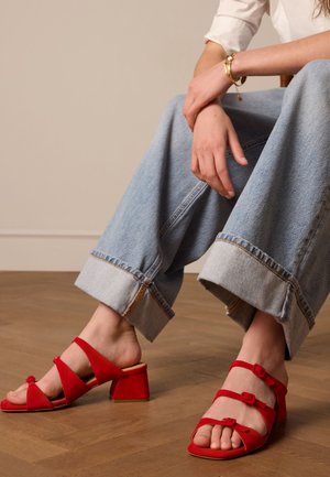 Rode suède blokhak sandalen met drie kruislings gespen en een bloemenaccent. Draag met lichtblauwe wijde denim jeans.