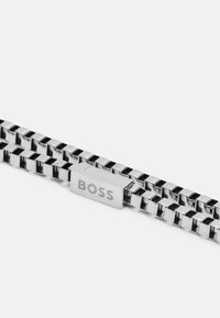 Bracelet en métal argenté avec des maillons entrelacés, doté d'un fermoir rectangulaire marqué "BOSS." Fini poli, texture uniforme et design élégant.