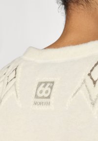 Prenda de forro polar blanca con patrones de diamantes cerca del escote. Presenta una etiqueta bordada gris que dice "66° NORTH". Textura suave.