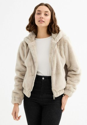 Chaqueta de piel sintética beige con capucha, cremallera frontal, puños elásticos y dobladillo; luciendo sobre una blusa blanca y combinada con jeans negros de cintura alta.