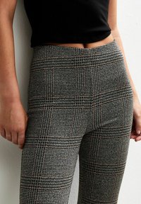 Gros plan sur une personne portant un pantalon à carreaux gris taille haute et un haut noir court, se tenant contre un mur blanc.