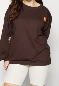 Felpa di cotone marrone con collo rotondo e maniche lunghe, caratterizzata da un accento grafico arancione sul petto. Indossata sopra pantaloni corti chiari.