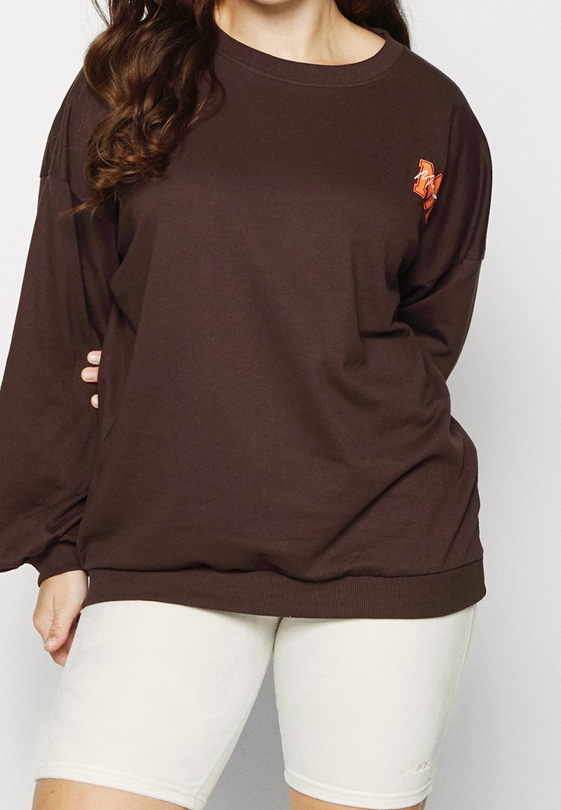 Felpa di cotone marrone con collo rotondo e maniche lunghe, caratterizzata da un accento grafico arancione sul petto. Indossata sopra pantaloni corti chiari.