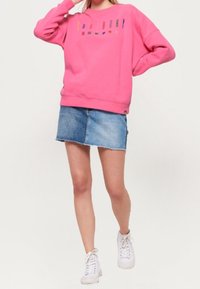 Pink sweatshirt with colorful graphic text, paired with a denim mini skirt. Model wears white sneakers, with a relaxed fit and crew neck design.
