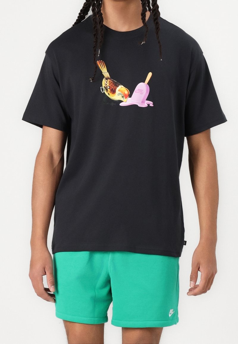 Camiseta de algodón negra con un gráfico colorido de un pájaro al lado de un helado de palo rosa. Combinada con pantalones cortos verdes, hechos de una tela suave.